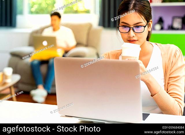 Asian working woman use computer notebook laptop with delivery coffee cup work from home in living room with social distance to couple in background while city...