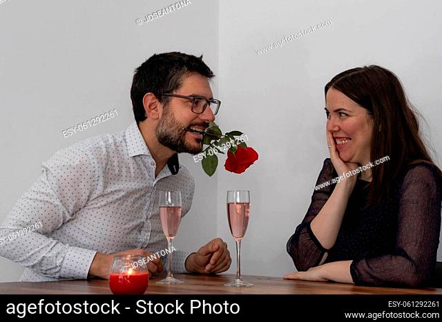 young man holding a rose in his mouth and his embarrassed smiling girlfriend with two glasses of champagne and candle