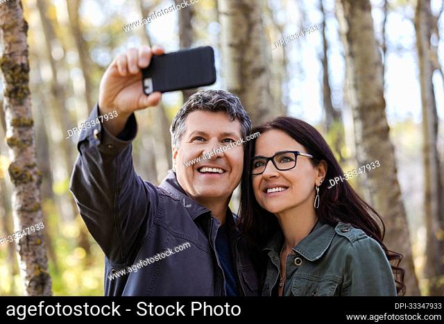 Mature couple taking a selfie with their phone and enjoying each other's company while taking a walk in the woods in autumn; St Albert, Alberta, Canada