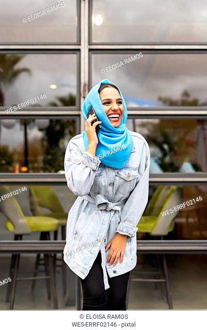 Young laughing woman wearing turquoise hijab and phoning in front of a cafe