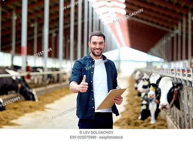 agriculture industry, farming, people and animal husbandry concept - happy smiling young man or farmer with clipboard and cows in cowshed on dairy farm showing...