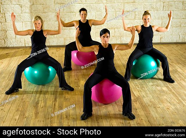 A group of women making exercise sitting on big balls with upraised hands. They're looking at camera. Front view