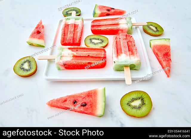 Creative pattern from different pieces of fruit and berry color ice cream on a stick in the shape of a square in a white plate on a gray marble background