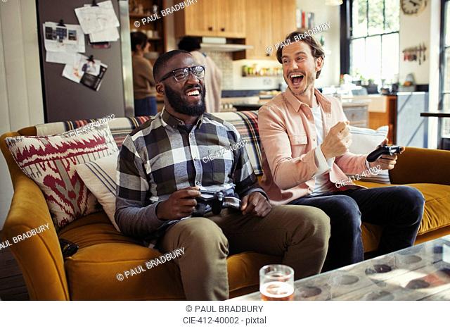 Laughing men friends playing video game on living room sofa