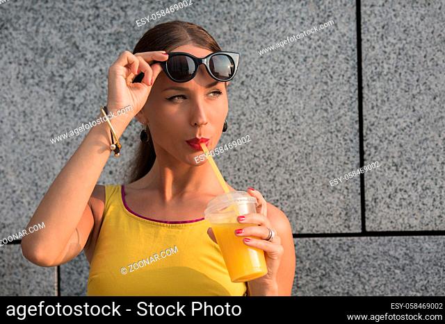 Fashion girl with red lips drinking orange cockrail against brick wall. Brown-haired beautiful lady in little yellow dress looking away
