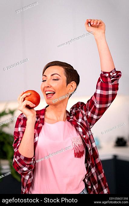 Holding an apple in hand happy woman dancing at kitchen with a sexy eye contact on camera. Charming housewife wearing plaid shirt with a short hair while...