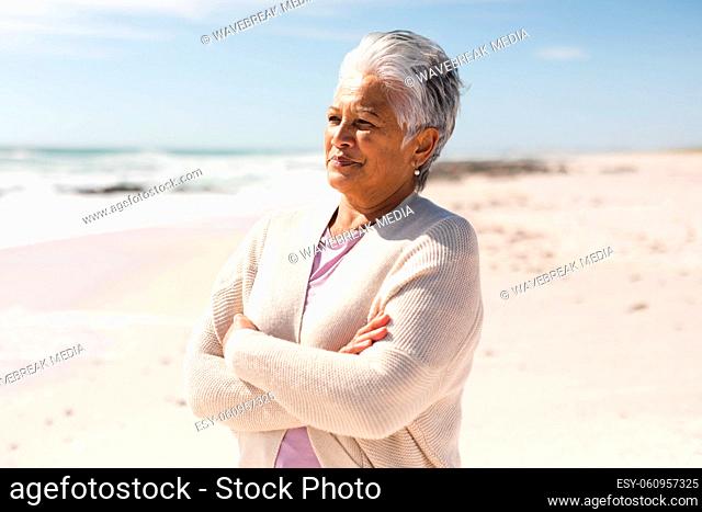 Thoughtful senior biracial woman with arms crossed looking away at beach against sky on sunny day