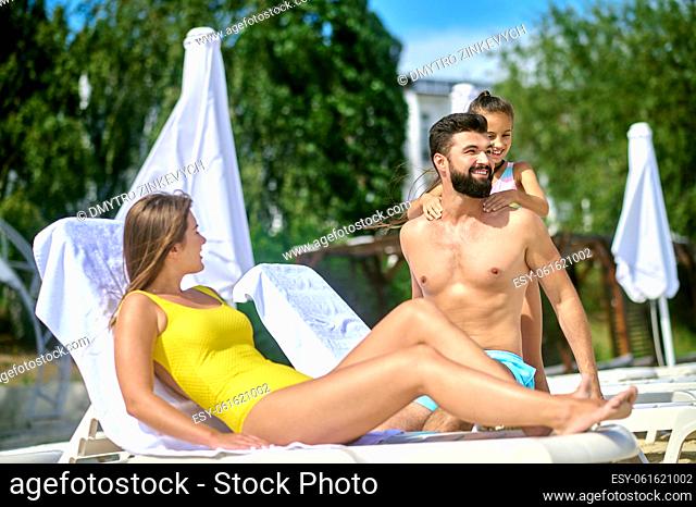 Woman in a swimming suit lying on the deckchair while looking at her pleased husband and cheerful daughter