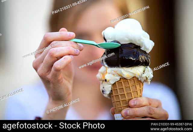 31 March 2021, North Rhine-Westphalia, Zons: A young woman eats her large ice cream in a cone with a spoon. Meteorologists predict spring-like weather for today...
