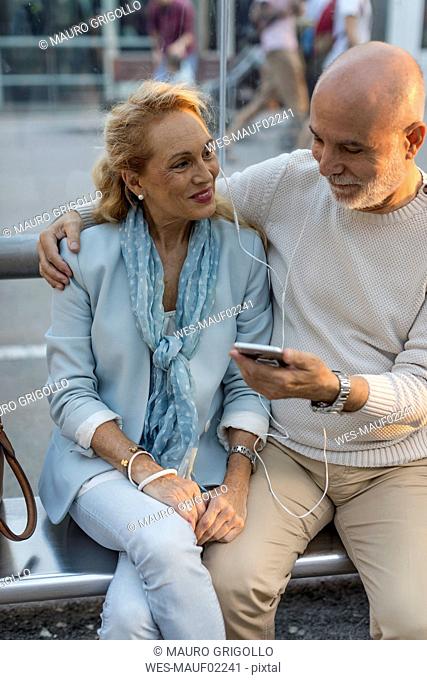Spain, Barcelona, senior couple sitting at tram stop in the city sharing smartphone with earbuds