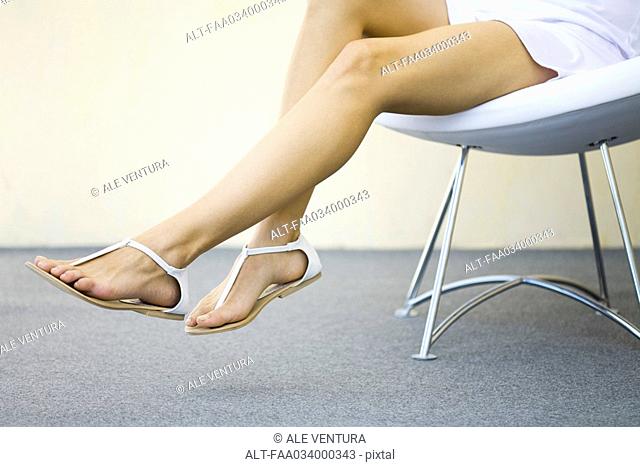 Woman sitting in chair with legs dangling, wearing sandals, cropped view