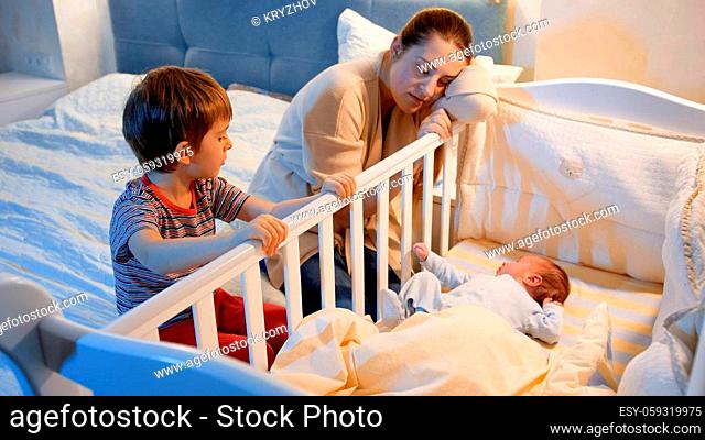Little boy with tired moteher rocking crib of newborn baby boy in bedroom at night. Children helping parents. Motherhood and sleepless nights