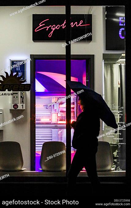 Stockholm, Sweden A woman with an umbrella walks by a tanning salon in Liljeholmen at night