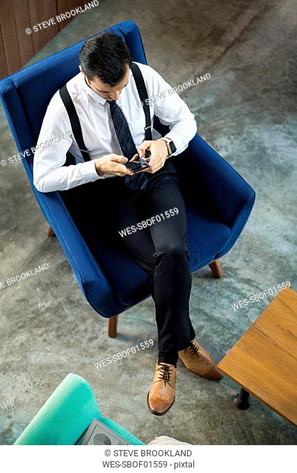 Top view of businessman sitting in blue armchair using smartphone