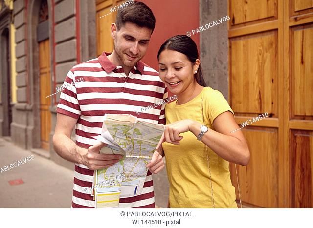 Young couple studying a map and pointing at it while smiling happily and dressed casually in t-shirts with old buildings behind them