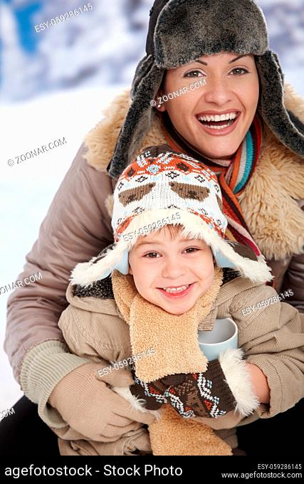 Portrait of happy mother and child holding cup of hot tea in snow on a cold winter day laughing, smiling