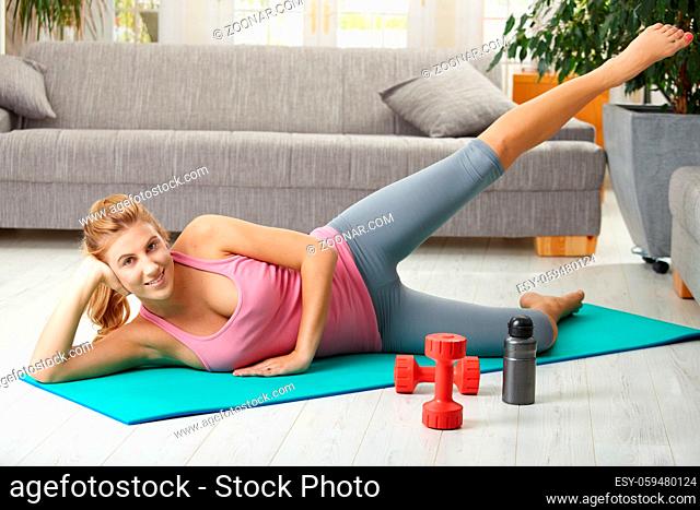 Young woman doing exercises lying on fitness mat in living room, smiling