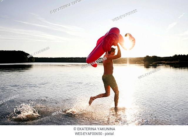 Young man running with flamingo pool float into a lake