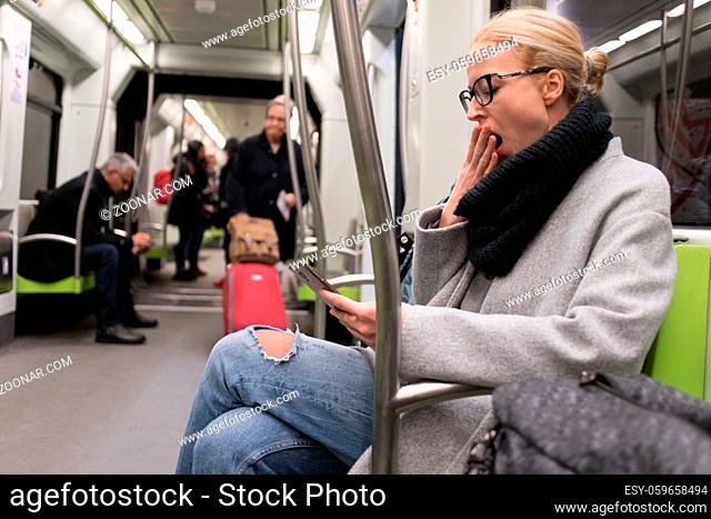 Beautiful blonde caucasian woman wearing winter coat and scarf yawning while reading on the phone, traveling by metro. Public transport