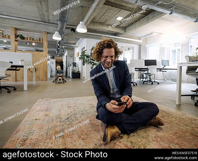 Businessman sitting on carpet in office using cell phone with son in background