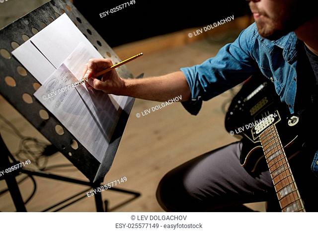 people, art and entertainment concept - man with guitar writing notes to music book at studio