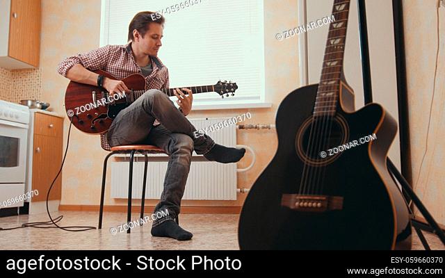 Young man musician composes music on the guitar and play in the kitchen, other musical instrument in the foreground, sitting