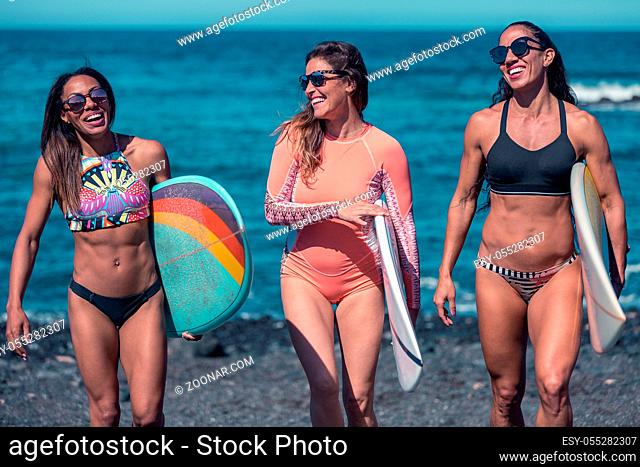 Group of cheerful diverse athletic women carrying surfboards and walking on seashore of ocean in sunlight