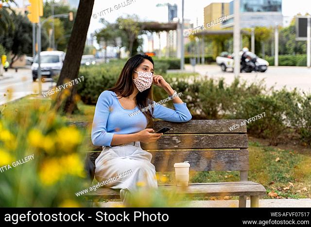 Contemplating woman looking away with smart phone while sitting on bench in city during COVID-19