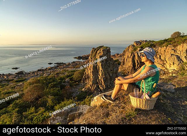 Woman sitting on a cliff in sunset having a picnic basket with wine, glasses and bread, nice warm evening light, Hovs hallar, Skåne, Sweden