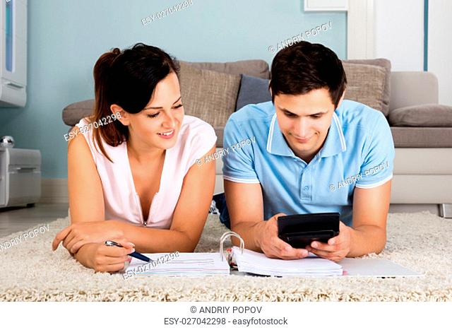 Happy Couple Lying On Carpet Calculating Bills Together At Home