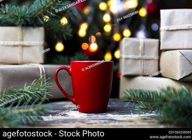 Delicious fresh festive morning coffee in a red cup on the rustic table with wrapped gifts, Christmas tree lights and spruce branches in the background