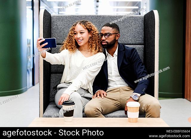 Business colleagues taking selfie sitting on seat at coffee break in coworking office