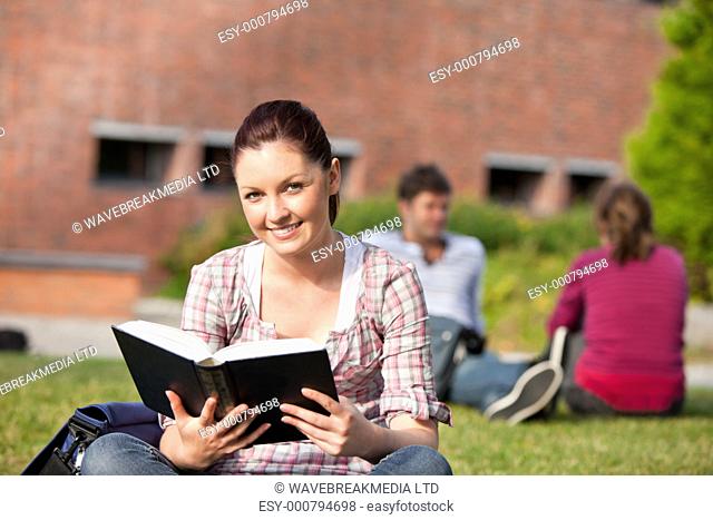 Delighted female student reading a book sitting on grass in the campus of her university