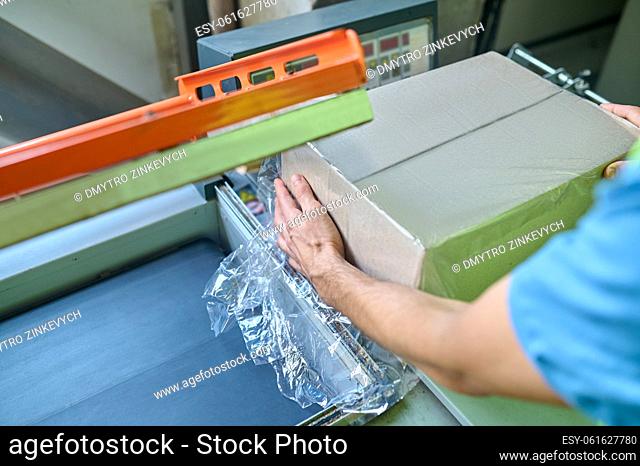 Cropped photo of a warehouse worker putting the cardboard box into the film wrapping machine