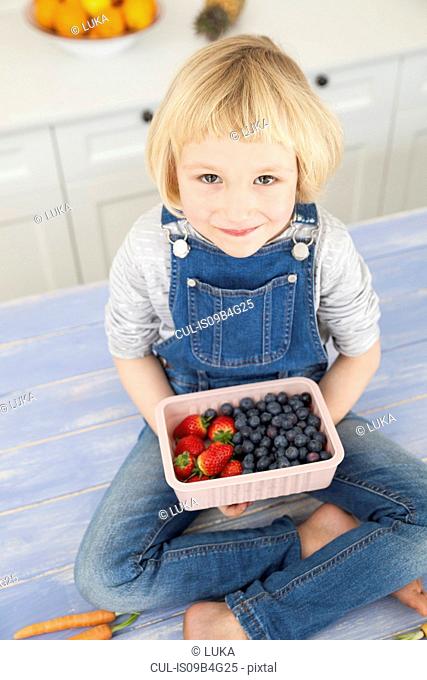 Portrait of cute girl holding punnet of blueberries and strawberries on kitchen counter