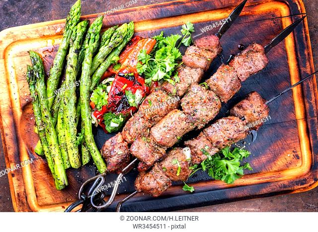 Traditional Russian shashlik on a barbecue skewer with green asparagus and paprika as top view on a burnt cutting board