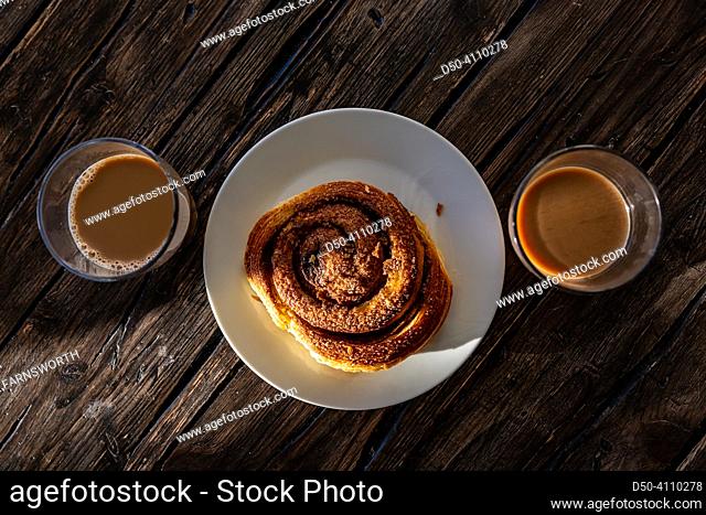 A sticky bun in the sunlight on a wooden table and two cups of coffee