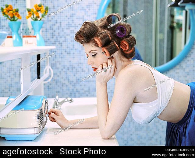 Portrait of a vintage styled woman turning on radio in a mid century bathroom, with hair curlers in her hair