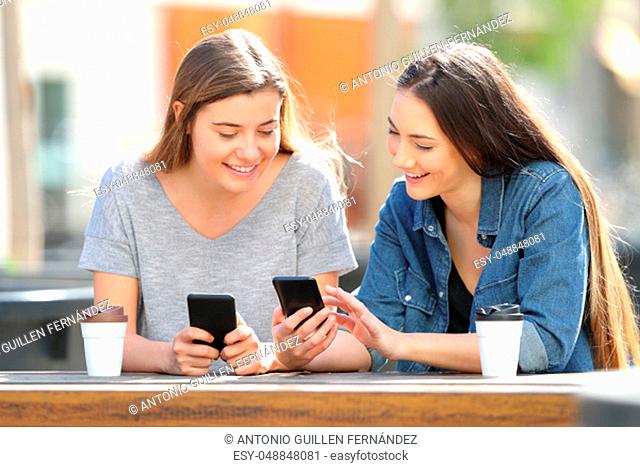 Two happy friends comparing their smart phones sitting in a park