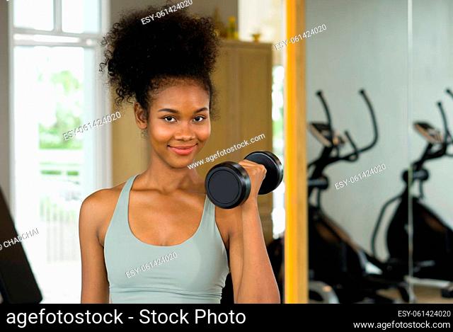 Young curly black hair woman enjoy holiday in fitness center. Lifting barbell with one hand while smiling
