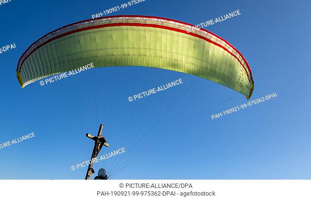 21 September 2019, Bavaria, Immenstadt: At midday on the mountain, the canopy of a paraglider taking off arches over the summit cross