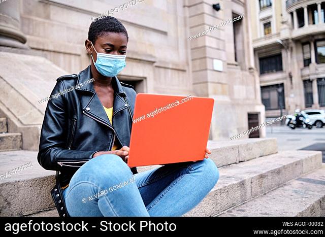 Young woman wearing protective face mask using laptop while sitting on steps in city