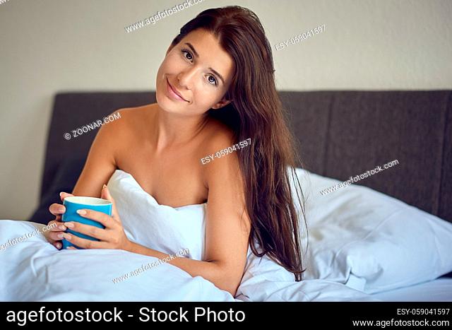 Beautiful young woman with long brown hair sitting in bed in the morning wrapped in white blanket with bare shoulders and holding blue coffee mug