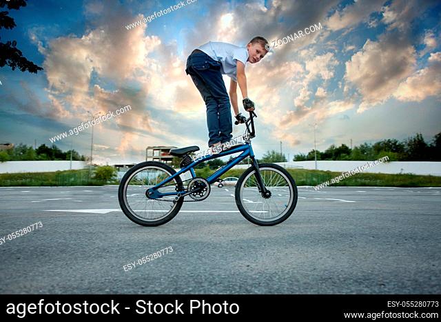 Close view of young sportive biker boy doing reckless tricks on his bike, looking into the camera. Cloudy sky background
