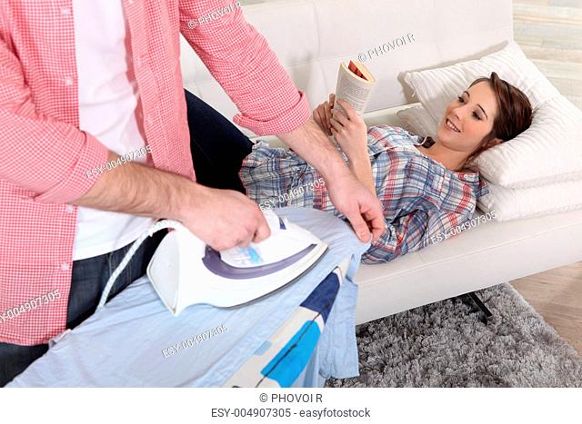 A man ironing while his girlfriend is reading on the sofa