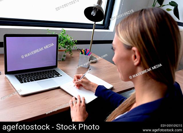 Caucasian businesswoman sitting at desk having video call writing notes using laptop with copy space