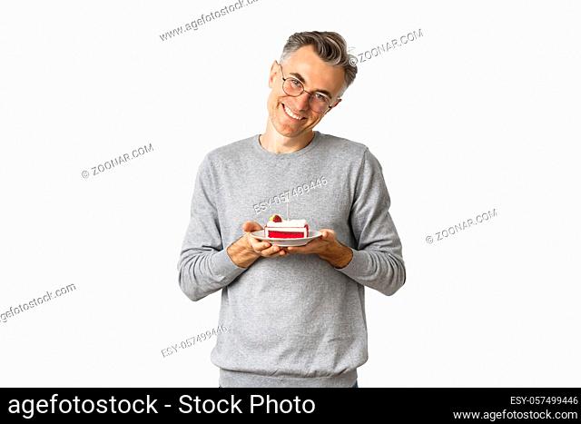 Portrait of smiling middle-aged man, looking touched and happy, celebrating birthday, holding b-day cake and celebrating, standing over white background