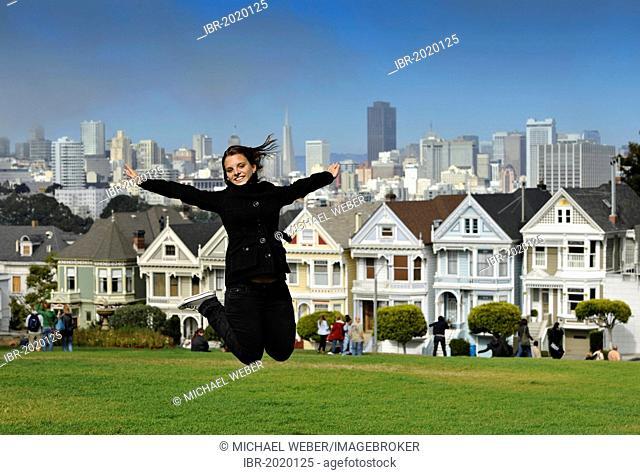 Young female tourist jumping in the air in front of the Painted Ladies, Victorian, multi-coloured wooden houses in front of the skyline with the Transamerica...