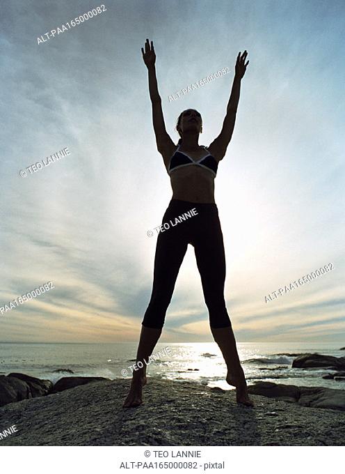 Young woman standing tiptoe, arms up, full length, on rocky shore, sky and seascape in background