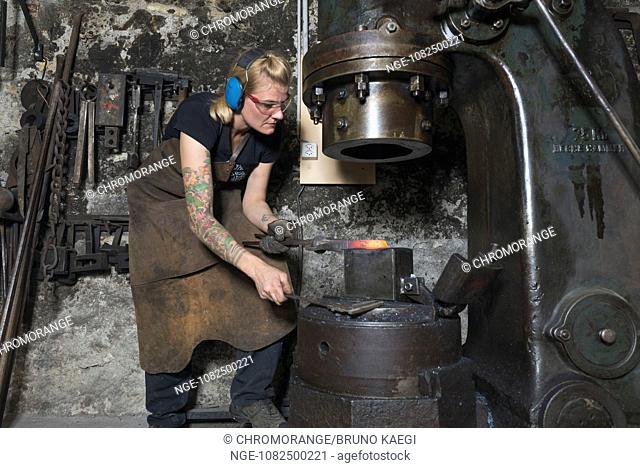 female blacksmith works a glowing piece of steel with steam hammer